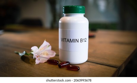 Close Shot Of White Plastic Bottle With The Words Vitamin B12 Written Across The Label With Capsules Set In The Foreground And An Out Of Focus Wooden Table Background.