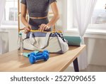 Close up shot of a woman hands packing a sports bag with gym equipment on a table at home. She is preparing for a training session or yoga workout, ensuring all fitness essentials are ready.