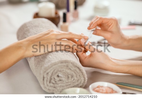 Close up shot of a woman in a nail salon receiving
manicure by beautician with metal nail file. Woman getting nail
manicure at spa centre. Beautician file nails to a customer in
luxury salon. 