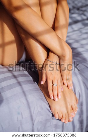 Close shot of womans hand with jewerly, bracelets and rings, hold legs. Woman sit on stripped gray blanket in bedroom at sunset light