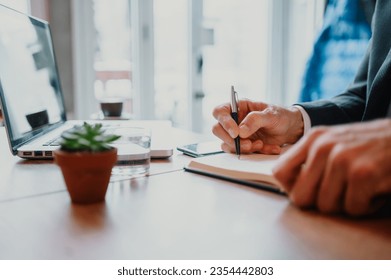 Close up shot of a wooden table with a small potted plant next to a glass of water and an open laptop. Focus on man's hands holding a pen and writing down in a notebook. Working remotely. Copy space. - Powered by Shutterstock