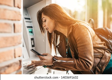 Close up shot of young adult woman. He typing pin code on keypad of ATM machine while using smart phone.
