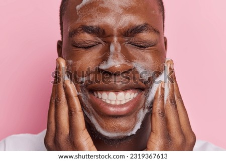 Close up shot of young African man exudes pure delight as he cleanses his face with soap pleased with condition of skin shows white teeth keeps eyes closed isolated over pink background. Daily hygiene