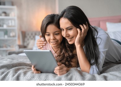 Close up shot of young attractive woman that hug her little daughter while lying on bed and holding using tablet. Mother and child look at the tablet screen in bedroom - Powered by Shutterstock