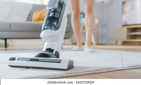 Close Up Shot of a Young Beautiful Woman in Jeans Shirt and Shorts Vacuum Cleaning a Carpet in a Bright Cozy Room at Home. She Uses a Modern Cordless Vacuum. She's Happy and Cheerful.