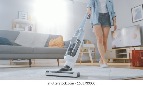 Close Up Shot of a Young Beautiful Woman in Jeans Shirt and Shorts Vacuum Cleaning a Carpet in a Bright Cozy Room at Home. She Uses a Modern Cordless Vacuum. She's Happy and Cheerful.