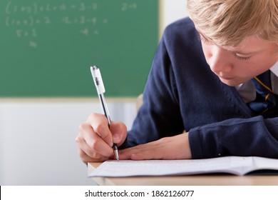 A close up shot of a young blonde boy in school uniform writing exam on his desk with black board in background. - Powered by Shutterstock