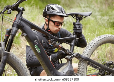 Close up shot of young Caucasian rider wearing protective gear adjusting seat of his battery-powered bicycle before ride, having concentrated look. Serious male repairing electric bike outdoors - Powered by Shutterstock