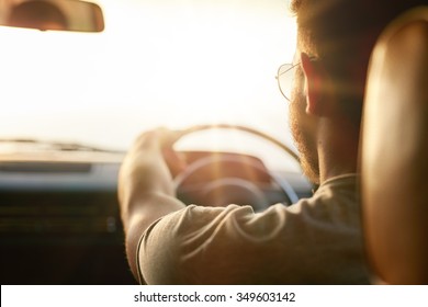 Close Up Shot Of Young Man Driving Car. Rear View Of Man On Road Trip On A Summer Day, With Sun Flare.