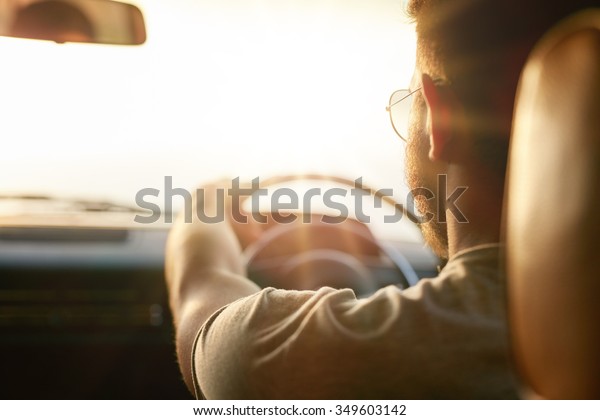 Close up shot of young man\
driving car. Rear view of man on road trip on a summer day, with\
sun flare.