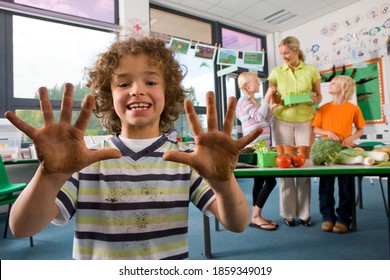 A Close Up Shot Of A Young Smiling Boy Showing His Dirty Muddy Hands To The Camera With Teacher And Other Students Standing In Background.