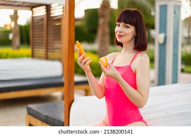 Close Up Shot Of A Young Woman Holding A Tube Of Sunscreen In Her Hands. Light-skinned Girl In A Pink Swimsuit Tanning Near Swimming Pool. Hot Summer Day. Sun Protection Skin Care Concept.