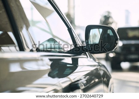 Close up showing side mirror and window of modern car reflecting dealership interior, sunlight highlighting vehicle surface, blurred background featuring another car in showroom