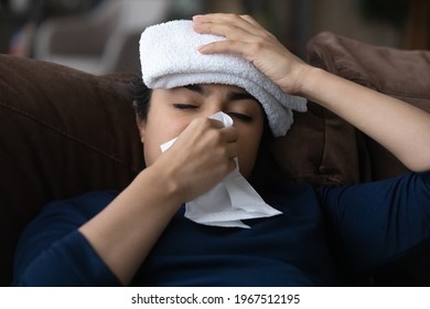 Close Up Sick Indian Woman Blowing Nose, Sitting On Couch At Home, Exhausted Young Female With Towel On Head Feeling Unhealthy And Unwell, Suffering From High Temperature, Catch Cold Or Flu