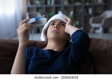 Close up sick Indian woman feeling unhealthy, holding thermometer, suffering from heat or fever, unhappy young female with towel on head leaning back on couch at home, cold or flu, health problem