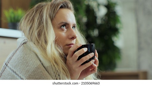 Close Up Of Sick Woman Sitting At Home In Blanket With Hot Tea Suffering From Fever Temperature Infection Flu Headache Cold. Young Person Drinking Tea, With Seasonal Virus Symptoms Feeling Unwell.