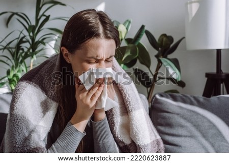 Close up of sick woman sitting on sofa freezing blowing running nose got fever caught cold sneezing in tissue, ill brunette girl covered with blanket, having influenza symptoms coughing at home