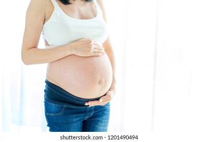 Close up side view of beautiful pregnant women standing near white curtain and touching on top and bottom of her belly. Expecting of future birth baby.  Healthy mommy concept.