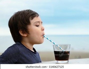 Close Up Side View Of Cute Kid Drinking Cold Drink In Coffee Shop By The Sea, Little Boy Drinking Soda With Straw. Child Drinking Cool Drink Soft Drink.