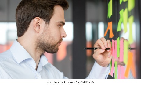 Close up side view focused businessman writing on colorful sticky notes attached on glass wall, entrepreneur create visual to-do list, future career business goals, daily aims, completed tasks concept