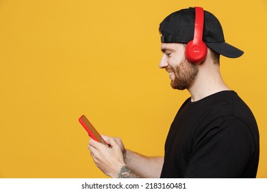 Close up side view fun young happy bearded tattooed man 20s he wearing casual black t-shirt cap headphones listen to music dance use mobile cell phone isolated on plain yellow wall background studio.