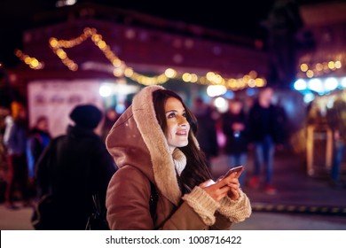 Close Up Side View Of Hooded Satisfied Cheerful Stylish Attractive Beautiful Young Happy Girl In Sweater And Jacket Looking At The Sky While Holding A Mobile On The Night City Street.