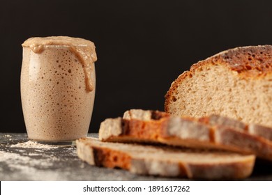 Close Up Side View Image Of A Freshly Baked Thick Crust Sourdough Artisan Bread With An Active Rising Sourdough Starter Culture In The Background. Rising Culture Is Overflowing The Glass Cup.