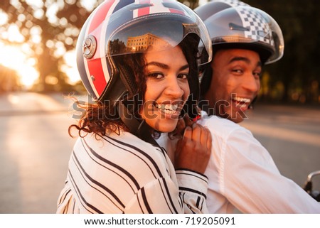 Close up side view image of happy african couple rides on modern motorbike outdoors and looking at the camera