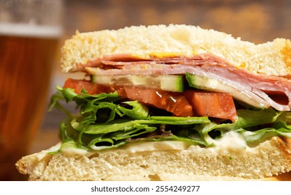 Close up side view isolated ham and salami sourdough bread sandwich. Mixed baby greens lettuce, mayo, mustard, havarti cheese, tomato, cucumber. Fresh delicious lunch. Glass cold beer beverage.