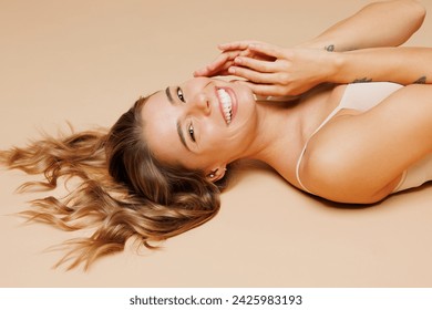 Close up side view relaxed satisfied happy young nice lady woman with slim body perfect skin wears nude top bra lingerie lay down isolated on plain pastel beige background. Lifestyle diet fit concept