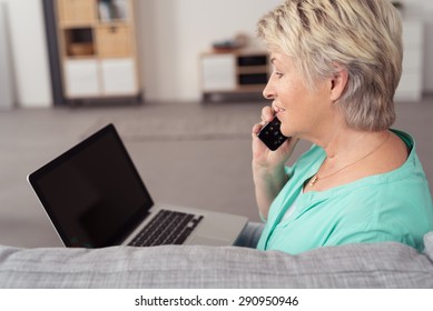 Close Up Side View Of A Senior Woman With Laptop Computer, Talking To Someone On Phone At The Living Room.