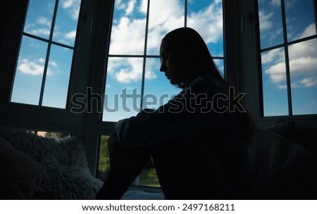 Close up side view of shadowed silhouette depressed woman while she sitting on windowsill with wide window on background. Sad, crisis, bad emotions, mental health, family violence.
