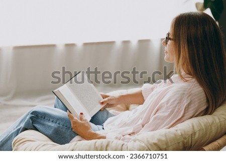Close up side view smart young woman wears casual clothes glasses sits in armchair read book stay at home hotel flat rest relax spend free spare time in living room indoor. Lifestyle lounge concept