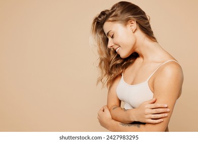 Close up side view smiling young nice lady woman with slim body perfect skin wear nude top bra lingerie stand hug herself isolated on plain pastel light beige background. Lifestyle diet fit concept