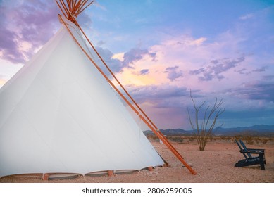 Close side view of a white teepee tent standing in open desert near Big Bend at colorful dusk. Canvas walls, wooden poles, purple clouds, blue sky, soft sunset glow, sandy ground, lone chair, sparse shrub, and distant mountains create a peaceful glamping scene with a quiet western feel. The image conveys remote travel, rustic lodging, natural texture, and calm outdoor living. It works well for adventure tourism, retreat promotions, desert lifestyle themes, and scenic vacation storytelling.
