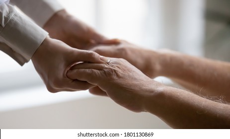 Close Up Side View Young Female Doctor Nurse Holding Wrinkled Hands Of Elderly Mature Patient, Helping Standing Up. Old 80s Man Involved In Rehabilitation Process With Professional Therapist Indoors.