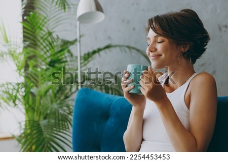 Close up side view young woman wear white tank shirt drink tea coffee sniff sit on blue sofa stay at home flat rest relax spend free spare time in living room indoors grey wall People lounge concept