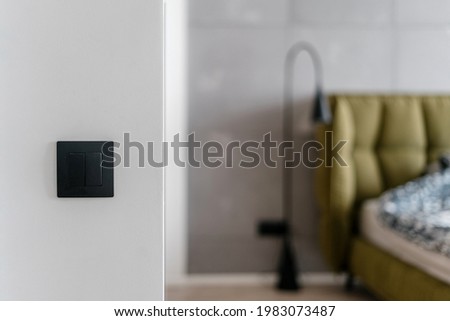 Close up of simple black matte light switch on white wall with bed and metal standing lamp in the blurred background, electrical system in stylish minimalistic loft style bedroom at home