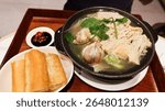 close up of a Singaporean white pepper soup Bak kut teh with pork rib, garlic, Flammulina filiformis in a black clay pot, spicy chili soy sauce and Chinese Fried Dough Sticks in a wooden tray