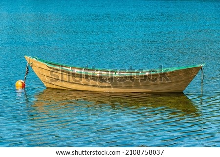 A close up of a single long yellow wooden dory with a red interior and green trim floating in blue still water. The row boat's reflections can be seen in the river along with strands of yellow grass.