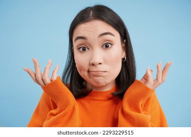 Close up of skeptical japanese girl, shrugs shoulders and looks confused, puzzled, stands over blue studio background. - Powered by Shutterstock