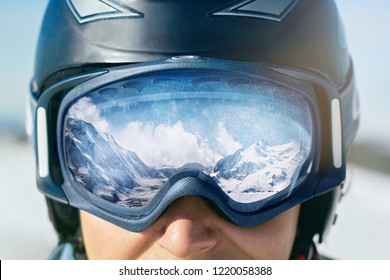 Close Up Of The Ski Goggles Of A Man With The Reflection Of Snowed Mountains.  A Mountain Range Reflected In The Ski Mask.  Man  On The Background Blue Sky. Wearing Ski Glasses. Winter Sports. 