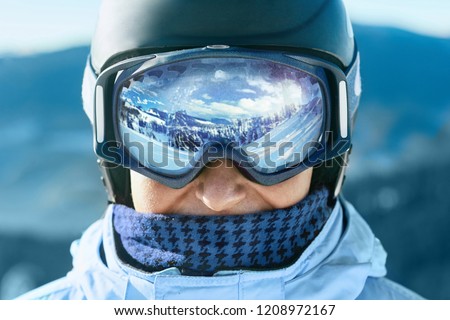 Close up of the ski goggles of a man with the reflection of snowed mountains.  A mountain range reflected in the ski mask.  Portrait of man at the ski resort on the background of mountains and sky