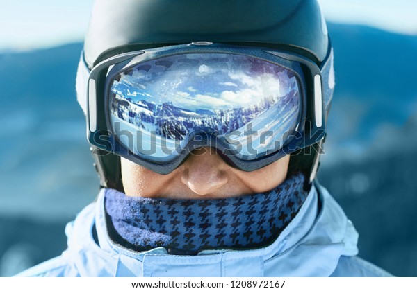 Close up of the ski goggles of a man with the\
reflection of snowed mountains.  A mountain range reflected in the\
ski mask.  Portrait of man at the ski resort on the background of\
mountains and sky