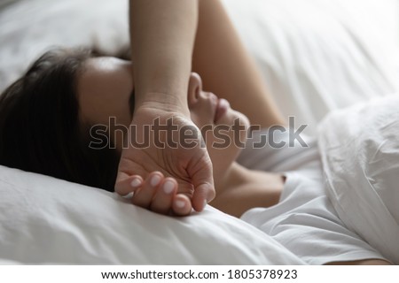 Close up sleepless young woman lying in bed, suffering from insomnia, tired exhausted female covering face with hand, trying to sleep, resting under white warm duvet on soft pillow