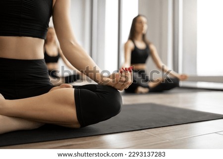 Close up of slim female in black top and yoga pant achieving lotus position with hands in mudra gesture on background of women in Padmasana posture. Advanced pose boasting physical benefits.