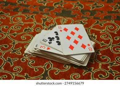 Close up of a small stack of playing cards with eight of diamonds visible on top placed on decorative red patterned fabric background, indoor tabletop still life scene.