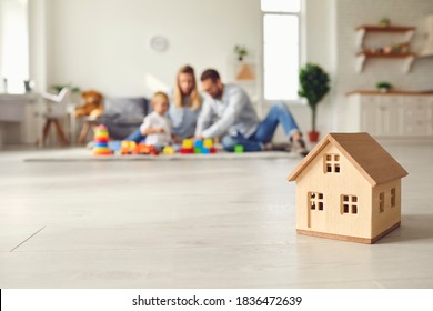 Close Up Of A Small Wooden House On The Floor In A Cozy Room On The Background Of A Young Family With A Child. House As A Symbol Of Buying A Property And Confidence In The Future. Real Estate Concept.