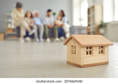 Close Up Of A Small Wooden House On The Background Of A Large Family Who Bought Their Own House. Concept Of Buying A House, Housing And Mortgage Lending. Blurred Background.