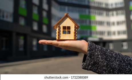 Close up of small wooden house in woman's hand on background of built house. Concept of purchasing new apartment.